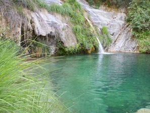A natural water body with a waterfall, surrounded by greenery and rocky terrain.