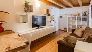 Uma sala de estar moderna com teto de madeira, uma TV de tela plana fixada em um armário branco, um sofá marrom e uma estante repleta de livros e objetos decorativos.