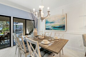 A dining area with a wooden table, white chairs, a chandelier, and a painting on the wall.