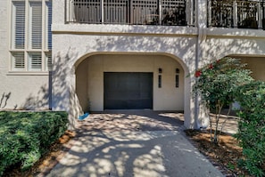 A two-story building with a garage entrance, a balcony with metal railings, and a small garden area with a tree and shrubbery.