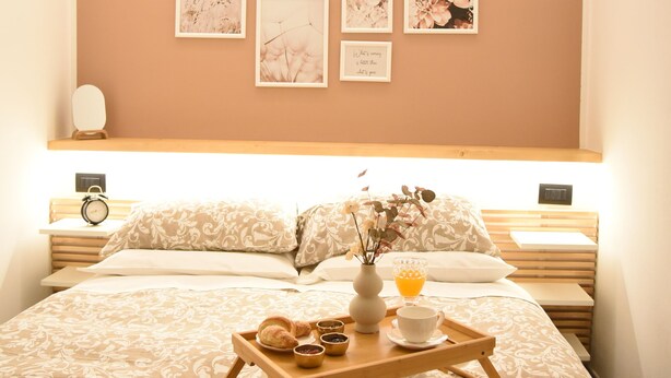 Een net opgemaakt bed met een tray ontbijtartikelen, een nachtkastje met een klok en lijstjes met kunstwerken aan de muur.