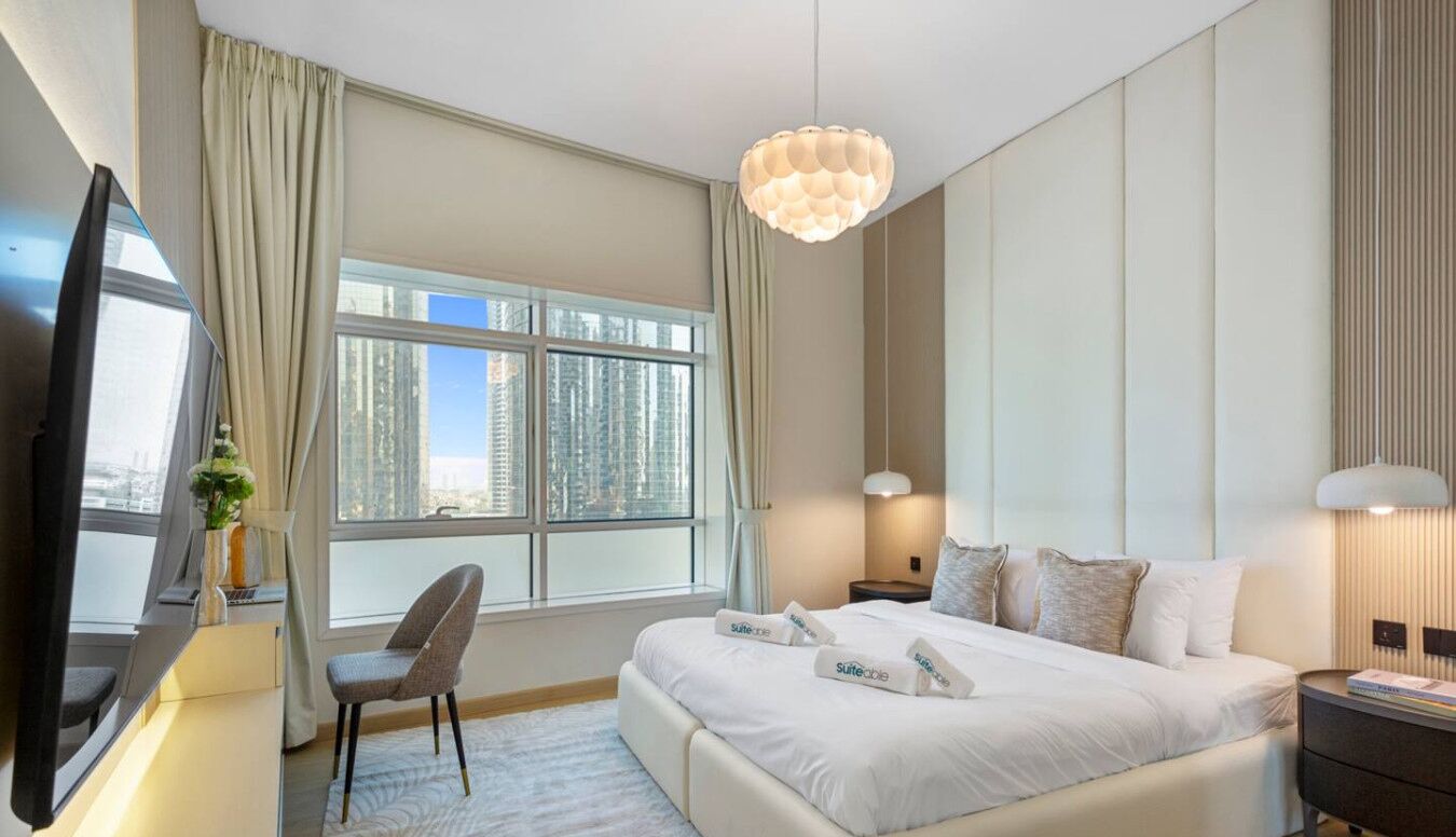 A hotel room with a large bed, a desk with a flat-screen TV, a chair, a bedside table, and a large window with a city view.