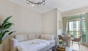 Moderni hotellihuone, jossa on sohva, ruokapöytä tuoleineen, ruukkukasvi ja suuri ikkuna, josta on näkymä rakennuksiin.