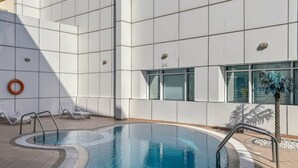 Un área de piscina interior moderna con agua azul cristalina, paredes de azulejos blancos y una palmera.