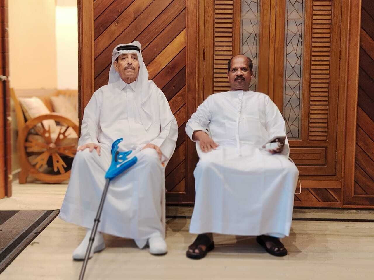 Two men in traditional white attire sitting in a room with wooden doors and a patterned wall.