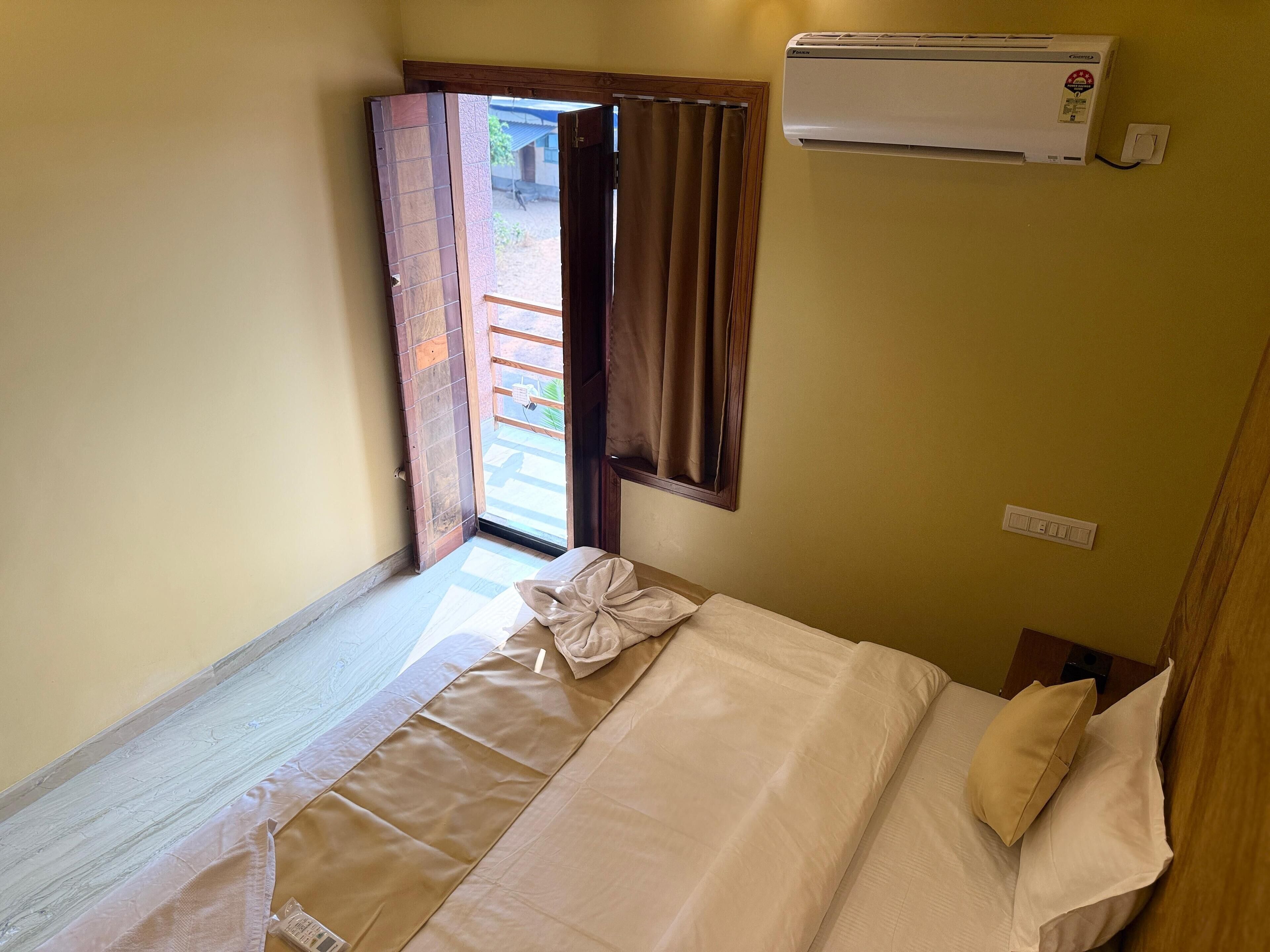 Una cama bien hecha con almohada y una manta doblada, una ventana abierta con cortinas y un aire acondicionado de pared.