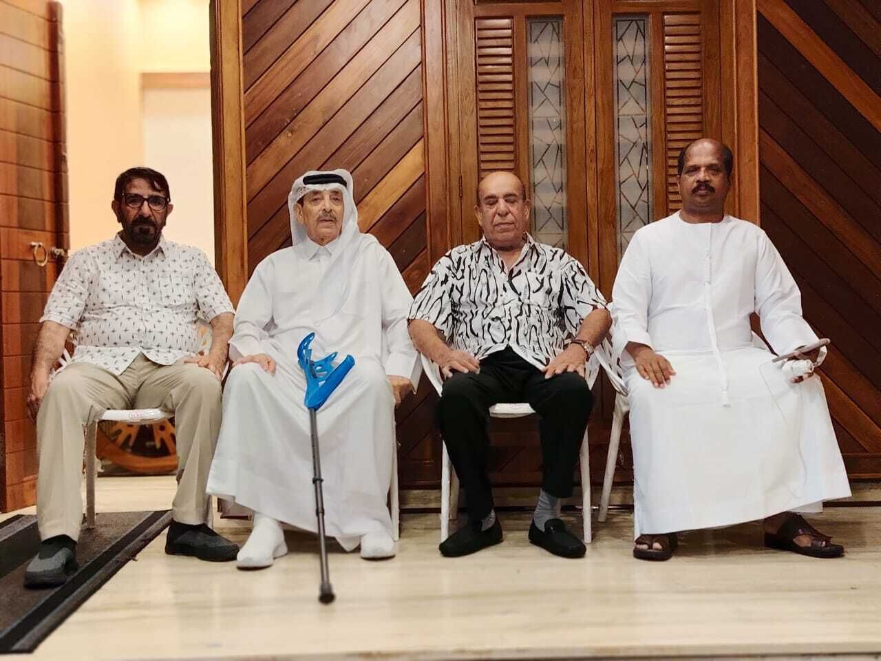 Four men seated on chairs in a room with wooden paneling and a door with glass panels.