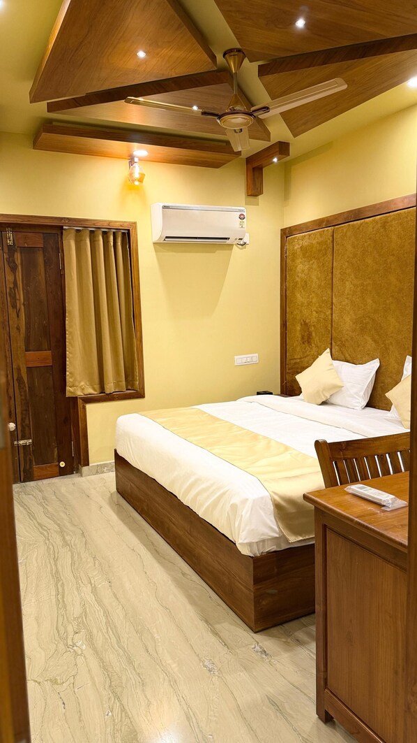 Quarto com cama de madeira, mesa de cabeceira, escrivaninha, ventoinha no teto e aparelho de ar condicionado.