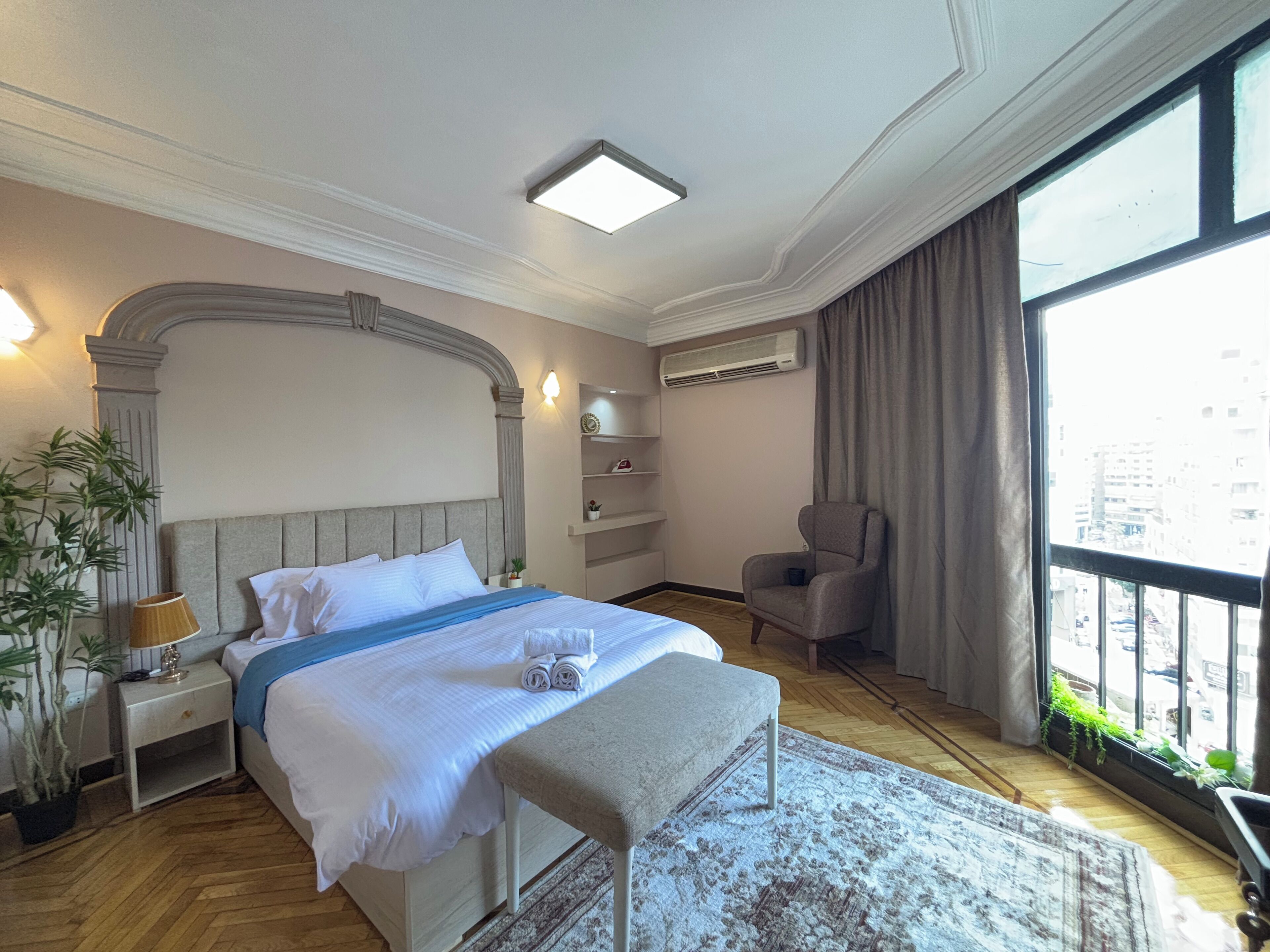 A hotel room with a large bed, a bench, a chair, a nightstand, a lamp, a potted plant, a shelf with decorative items, an air conditioning unit, and a large window with a view of buildings.