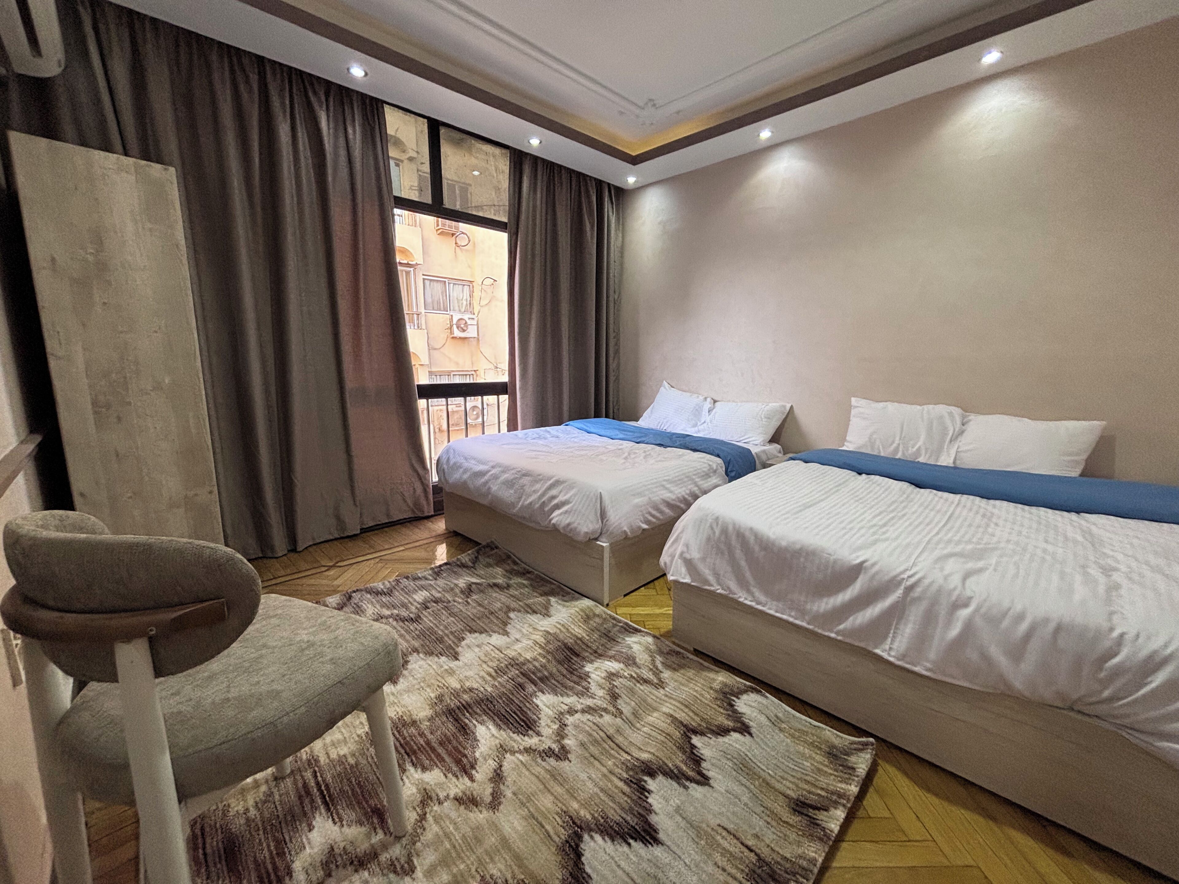 A hotel room with two beds, a chair, a patterned rug, and a view of the outside through a window with curtains.