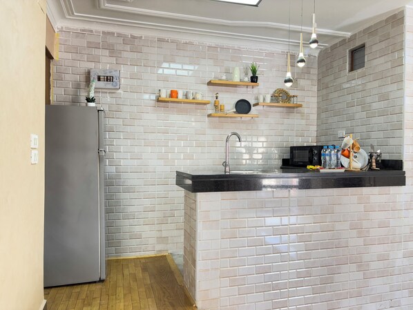 A kitchen with a grey refrigerator, a black microwave, and a black countertop. There are two wooden shelves above the sink, holding various items. - Joy Homes Mohandeseen - Families only (Giza)