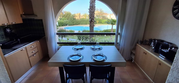 A kitchen with a dining table set for two, a view of a swimming pool, and a clock on the wall. - Apartment in a residential complex with two pools, 100 meters from the beach (Linguizzetta)