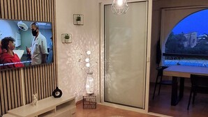 Un salón moderno con un televisor de pantalla plana, pared con paneles de madera, dos mesas redondas y un amplio ventanal con vistas al exterior.