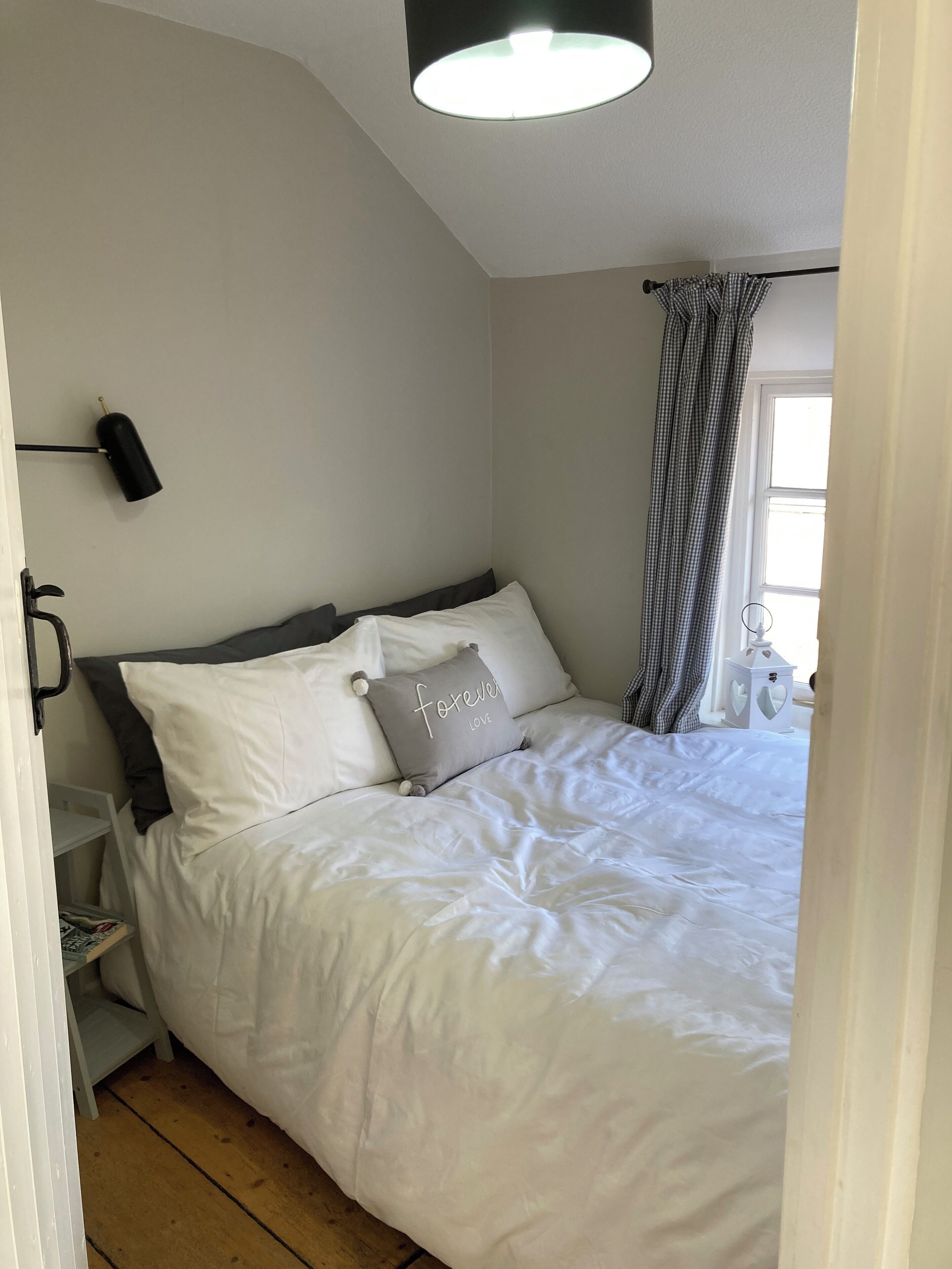 Quarto com cama, mesa de cabeceira, luminária, janela com cortinas e apoio para objetos pessoais.