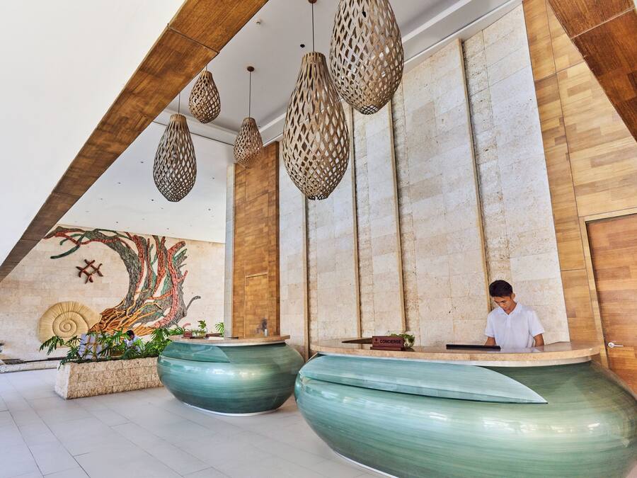 Un área de recepción con recepcionista, grandes macetas verdes, detalles de madera y un mural en la pared.
