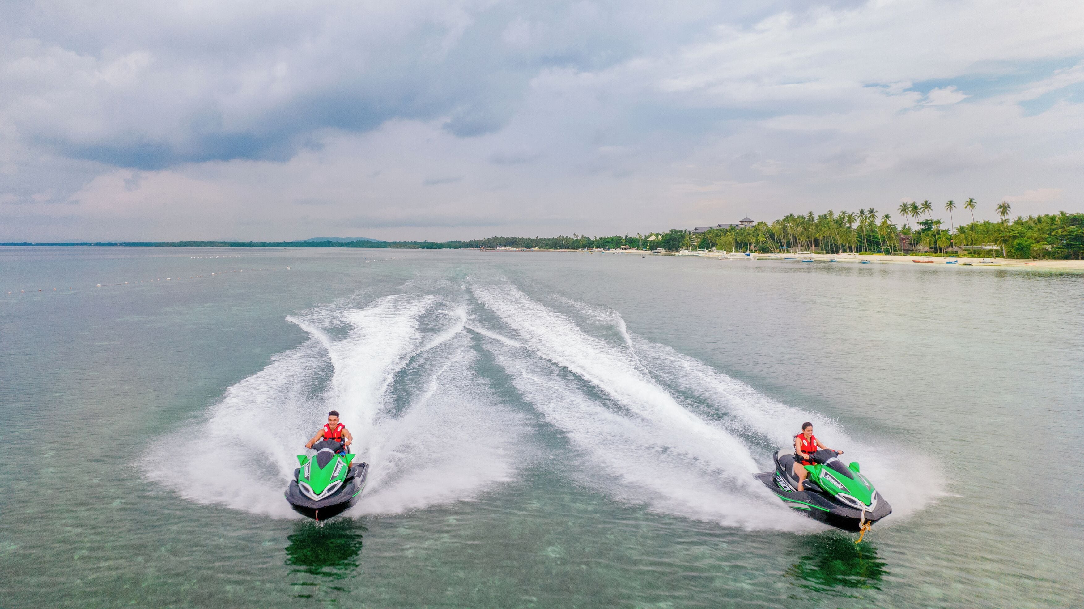 To jetski'er på roligt vand med en strand og palmer i baggrunden.