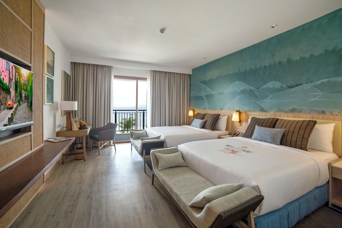A hotel room with two beds, a desk, a chair, a TV, and a large painting on the wall.