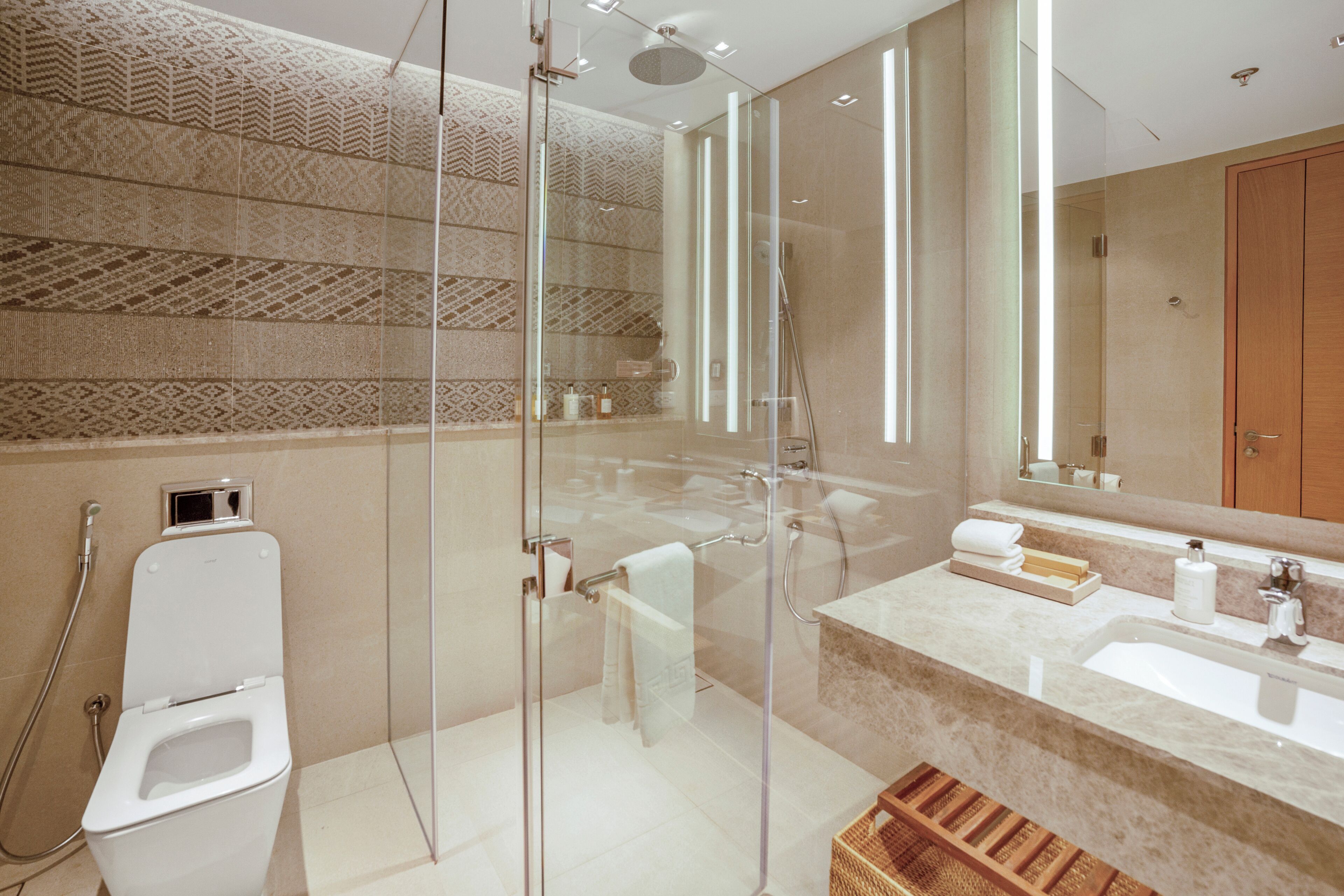 Un baño moderno con ducha de vidrio, inodoro blanco, lavamanos con encimera de mármol y una cesta de madera.