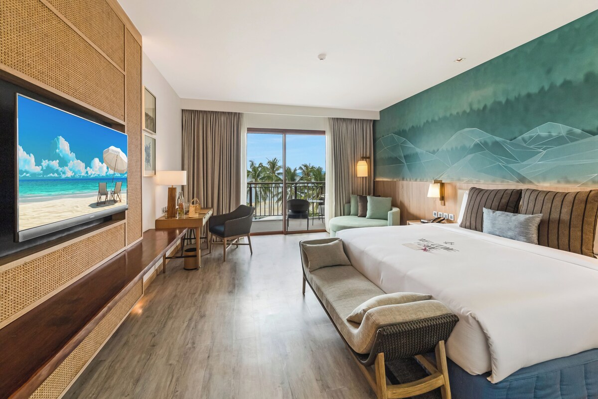 A hotel room with a large bed, a TV, a balcony with a view of palm trees, and a wall mural.