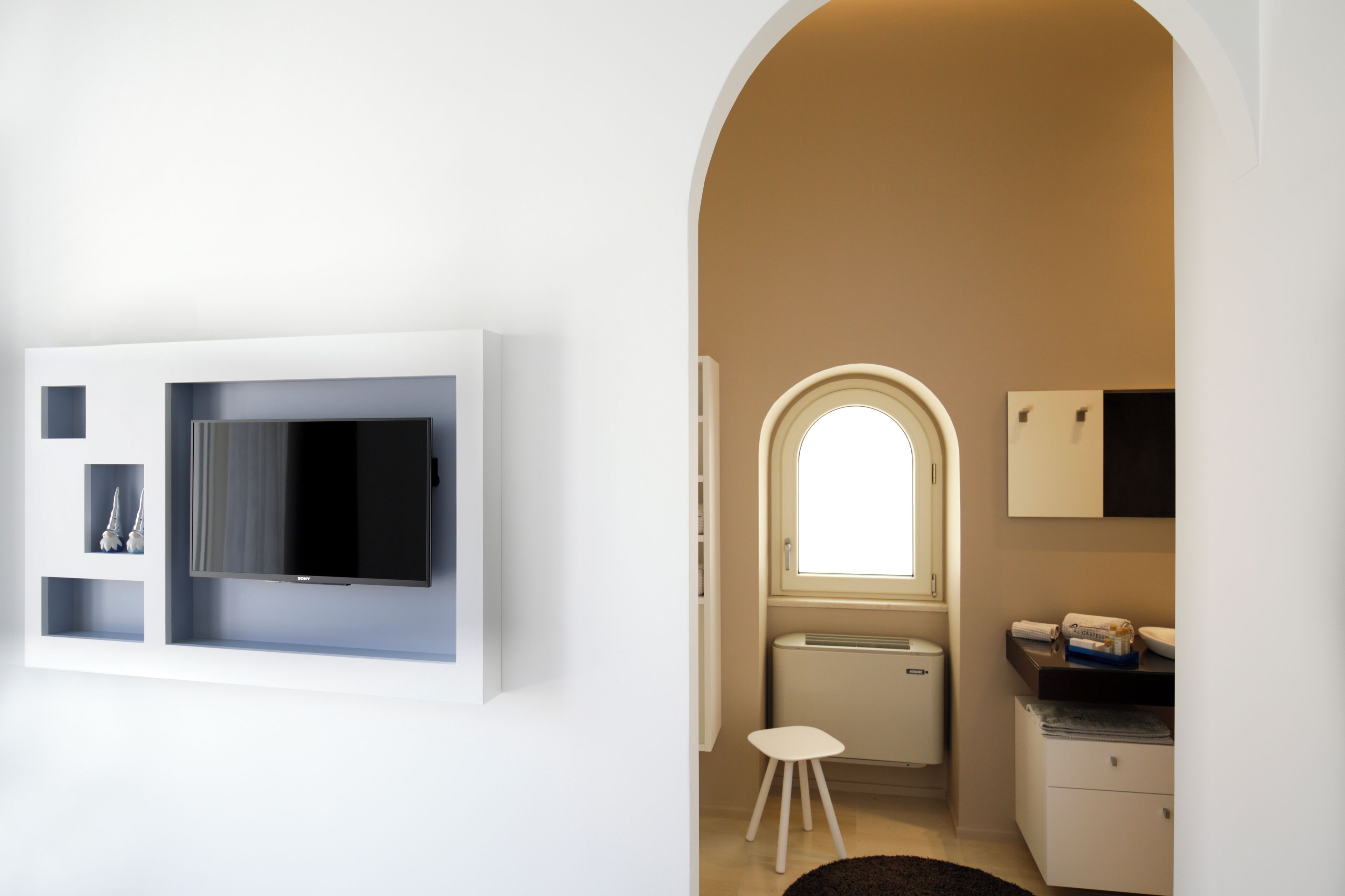 A modern room with a flat-screen TV mounted on the wall, a small white stool, a radiator, and a window with an arched top.