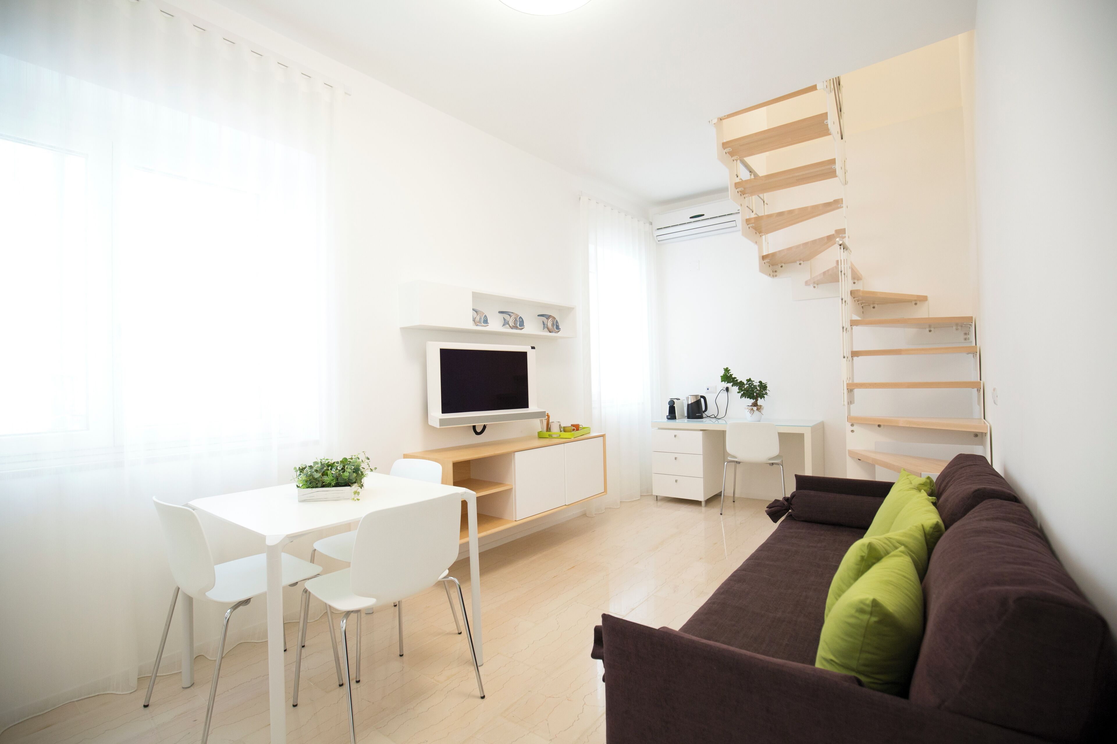 A modern, well-lit room with a dining table, white chairs, a brown sofa with green cushions, a TV, a desk with a computer, and a wooden spiral staircase.