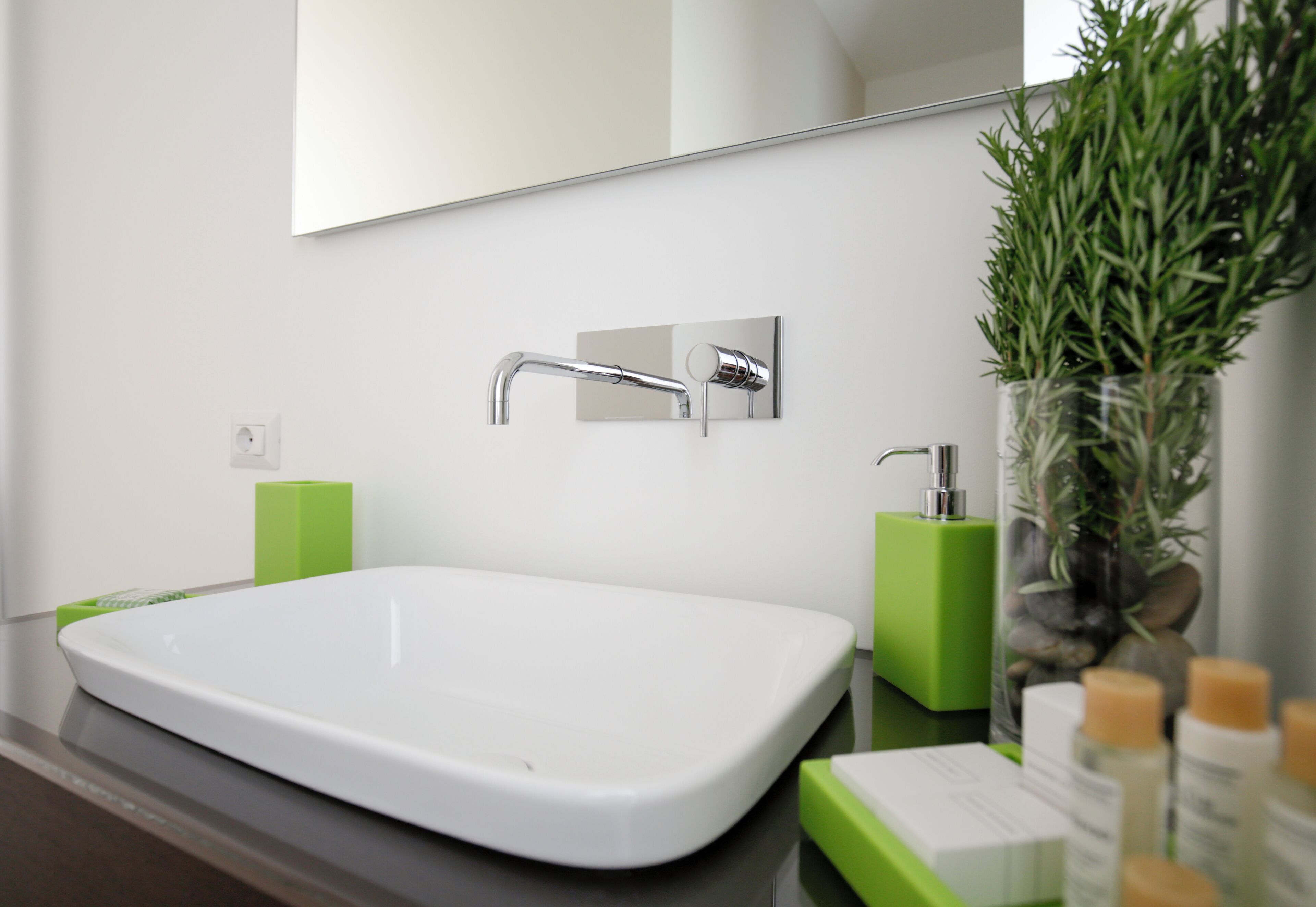 A modern bathroom with a white sink, green soap dispensers, and a potted plant.