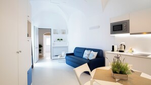 A modern, well-lit living area with a blue sofa, a dining table with a plant, and a kitchen with a microwave. - Al Castello Relais (Vieste)