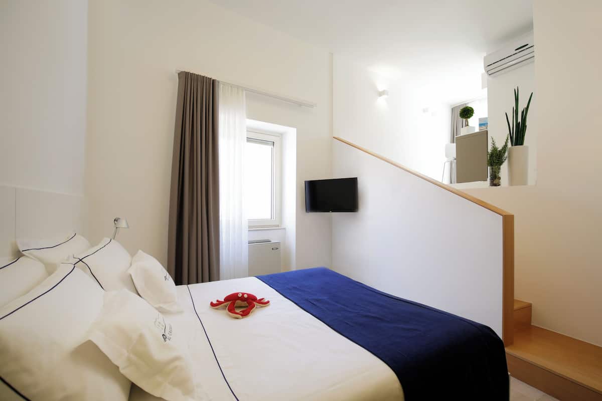 A hotel room with a bed, a flat-screen TV mounted on the wall, a window with curtains, a staircase, and a potted plant.