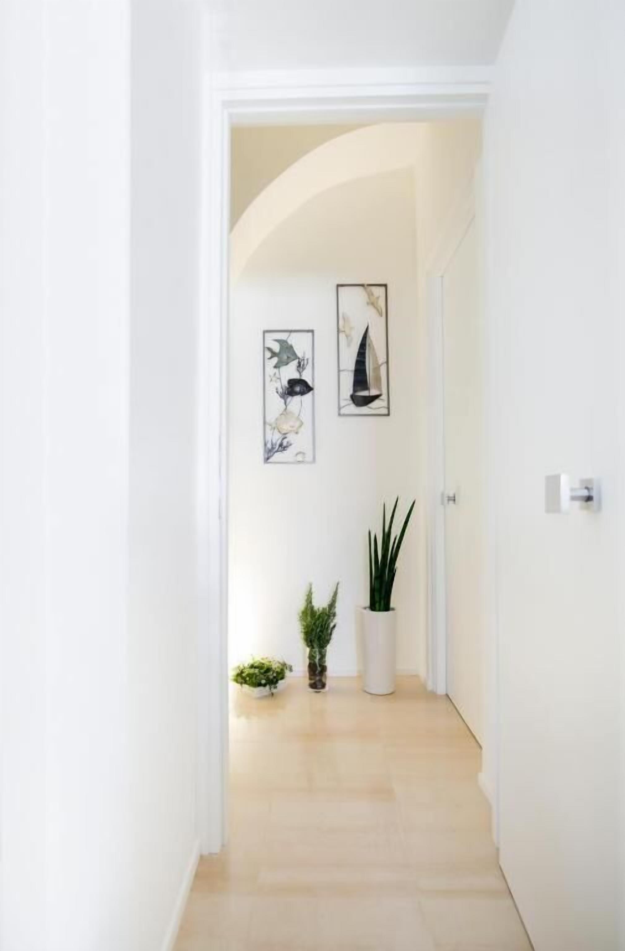 Un pasillo con una puerta blanca, dos cuadros enmarcados y plantas en macetas.
