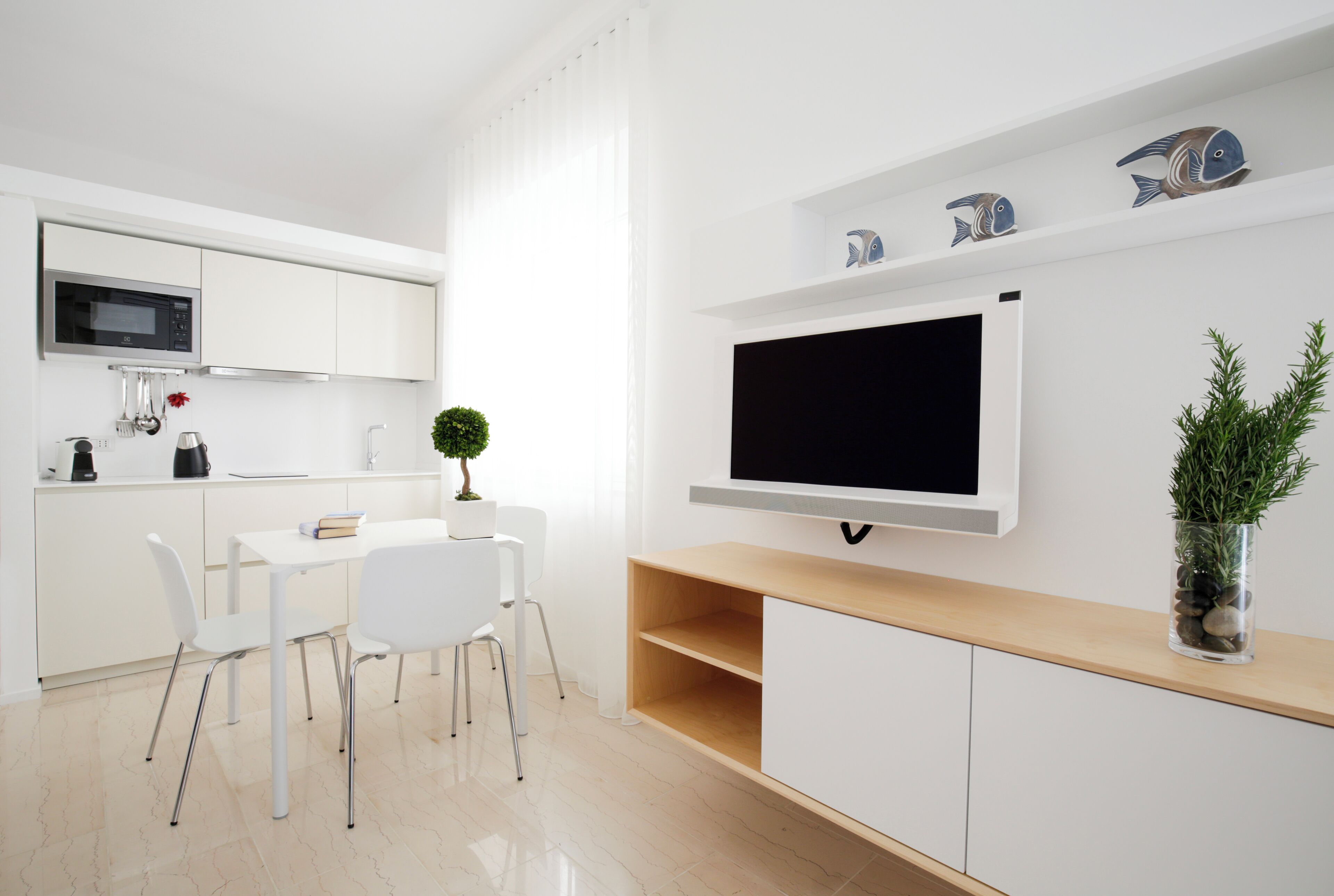 Une cuisine moderne avec des meubles blancs, un four à micro-ondes encastré, un coin repas avec des chaises blanches et un téléviseur fixé au mur.