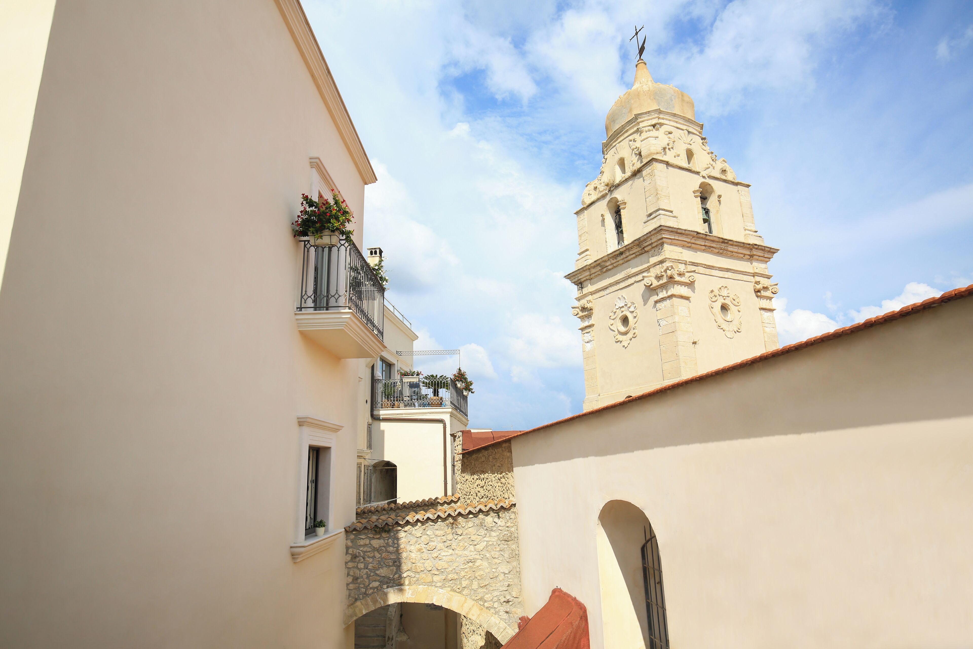 A narrow street with a building featuring a balcony with potted plants and a bell tower in the background.