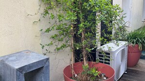 Una planta en maceta, dentro de una olla roja grande, un banco de metal y un equipo de aire acondicionado en una terraza de madera.