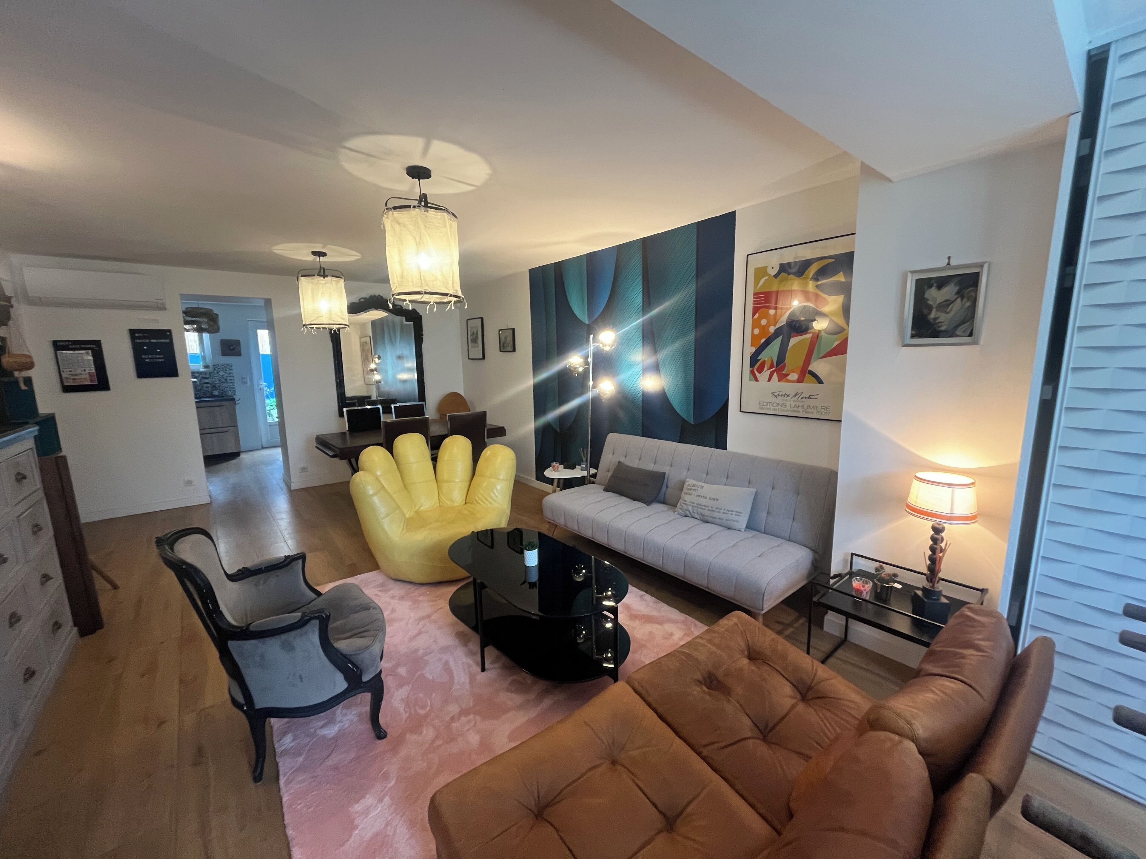 A modern living room with a sofa, armchair, and a coffee table. There is a dining area with a table and chairs, and a kitchen in the background.