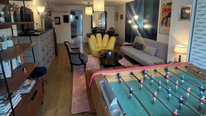 Una sala de estar acogedora con mesa de fútbol, un sofá, un sillón amarillo y un comedor con mesa y sillas.