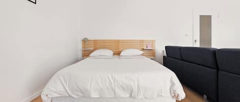 A bedroom with a wooden bed, white bedding, a dark sofa, and a framed picture on the wall.