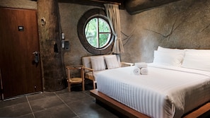 A room with a large bed, a round window, a wooden door, and a small table. - Lush Inn Phuket (Chalong)