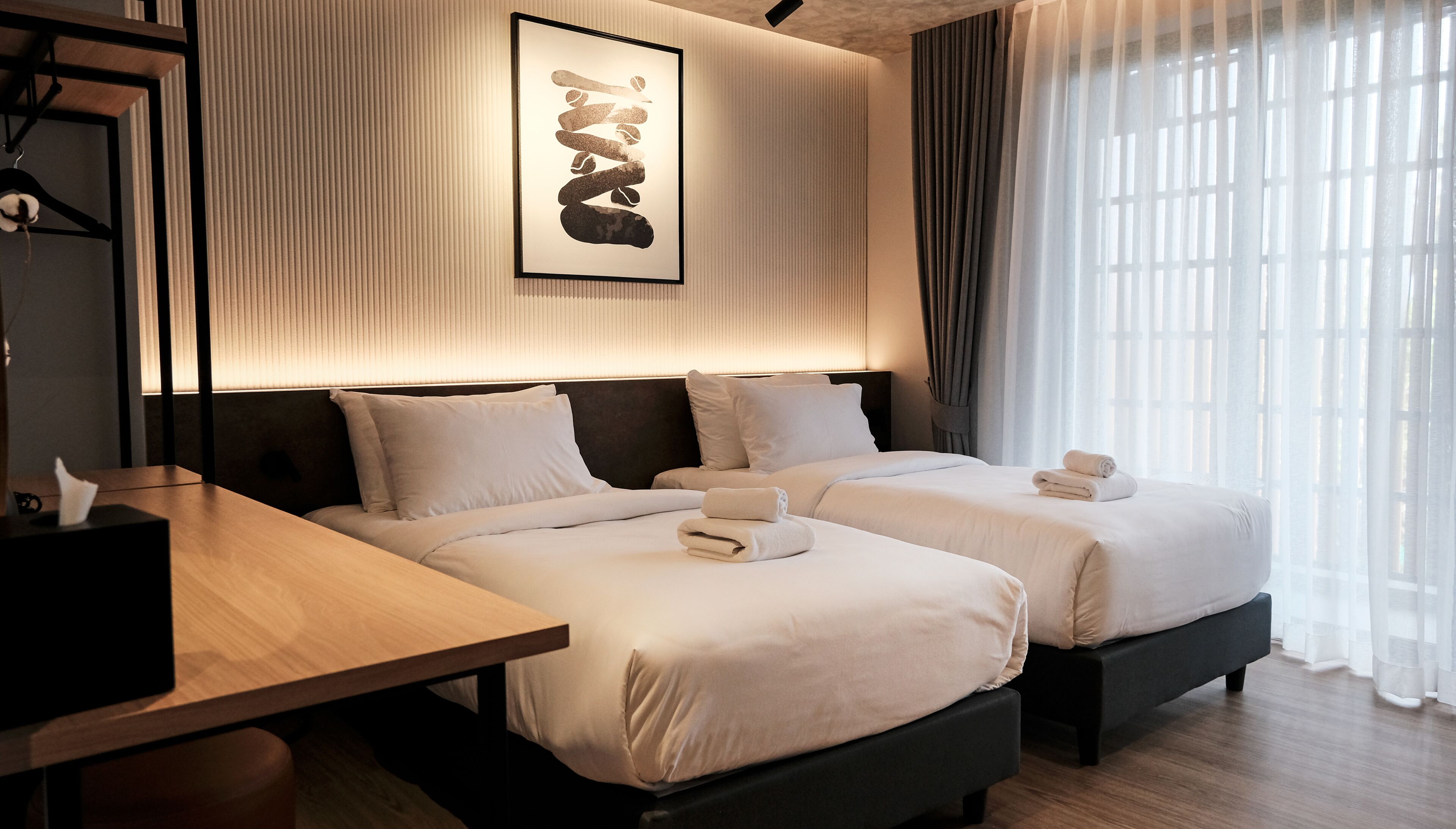 A hotel room with two beds, a bedside table, a wall-mounted artwork, and a window with curtains.