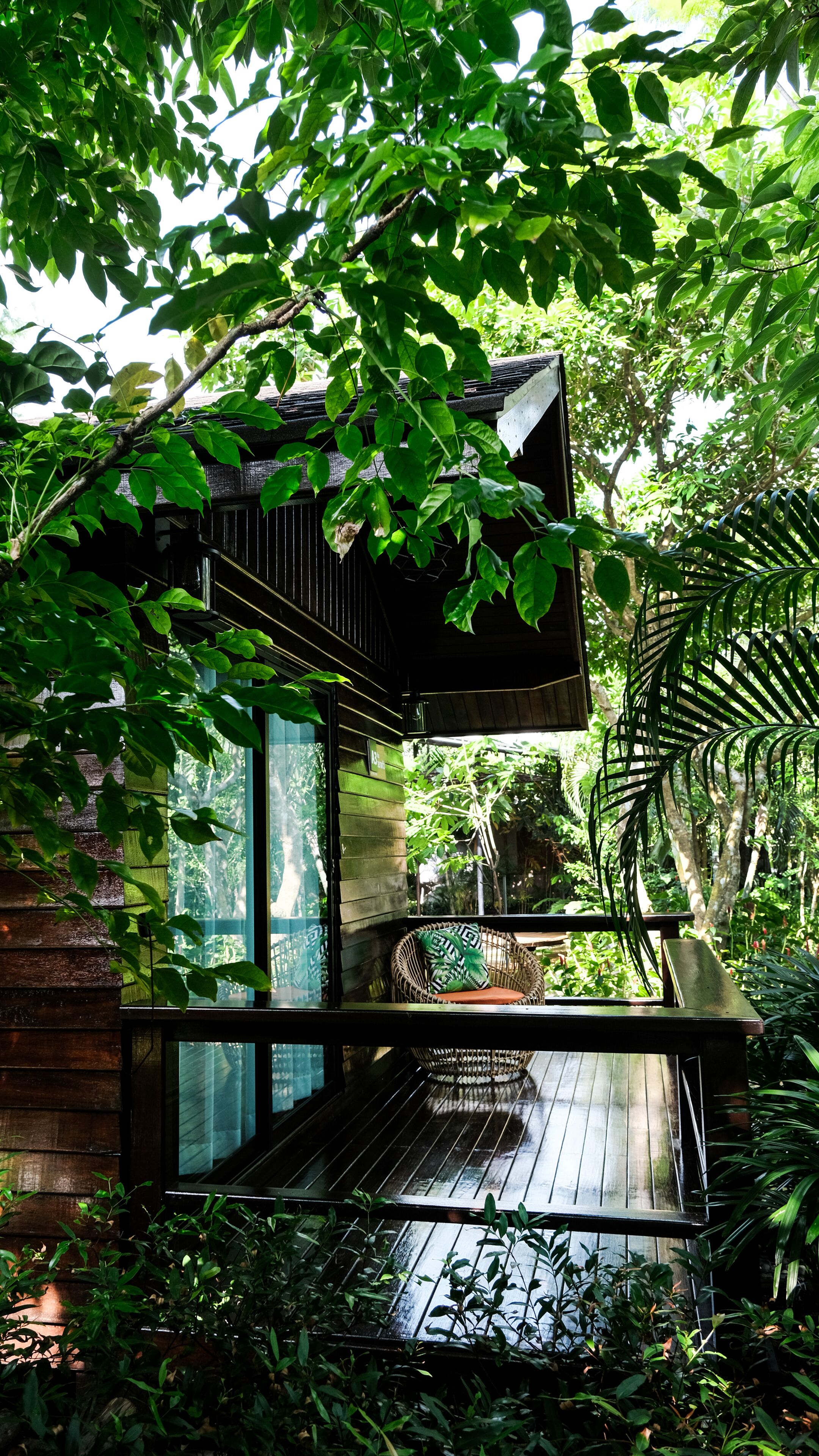A modern outdoor seating area with a wooden deck, surrounded by lush greenery and a hanging chair.