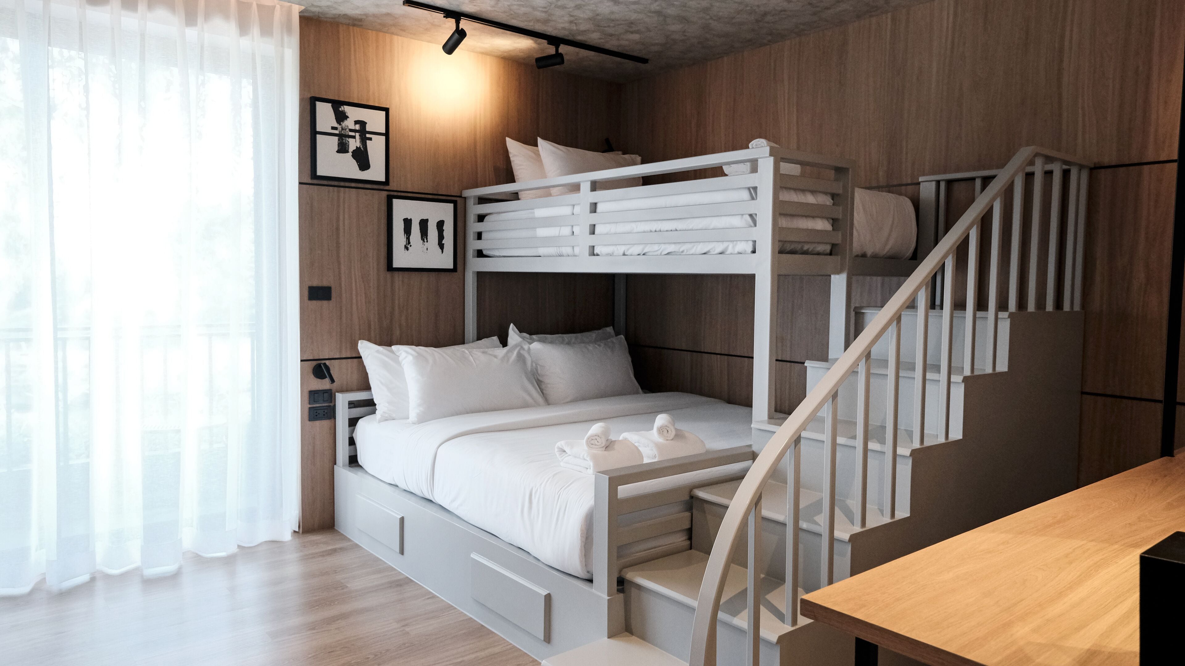 Ein Schlafraum mit Etagenbett, einer Holztreppe, einem Nachttisch und einem Fenster mit durchsichtigen Vorhängen.