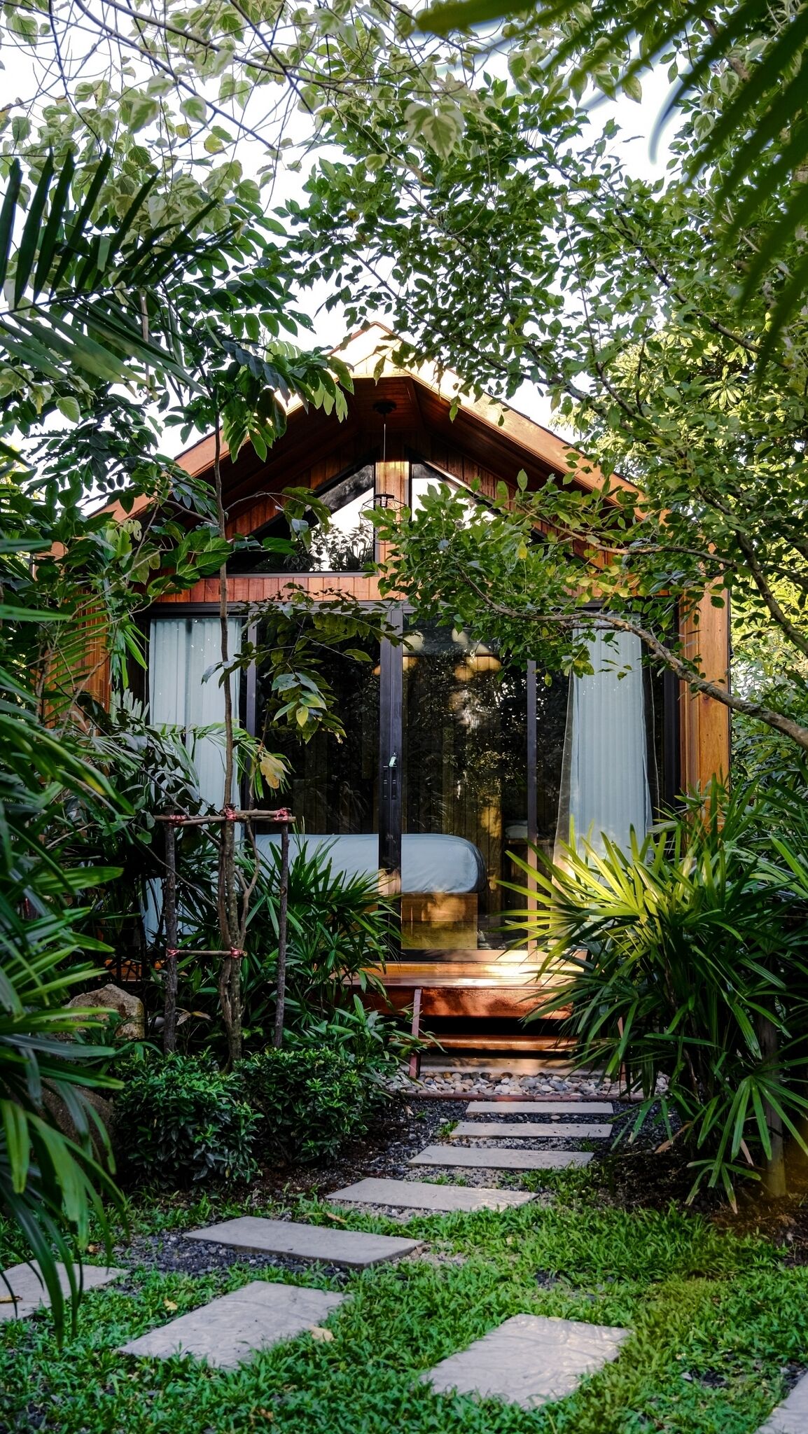 Una casa de madera con puerta de cristal, rodeada de abundante vegetación y un sendero de piedra que conduce a la entrada.