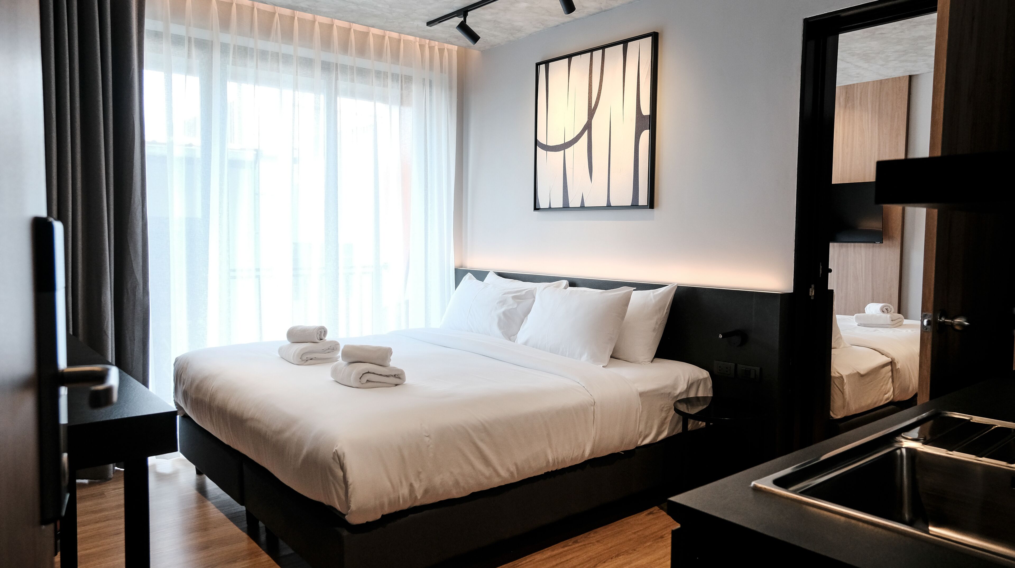 A modern hotel room with a large bed, a bedside table, a mirror, and a view of the city through sheer curtains.