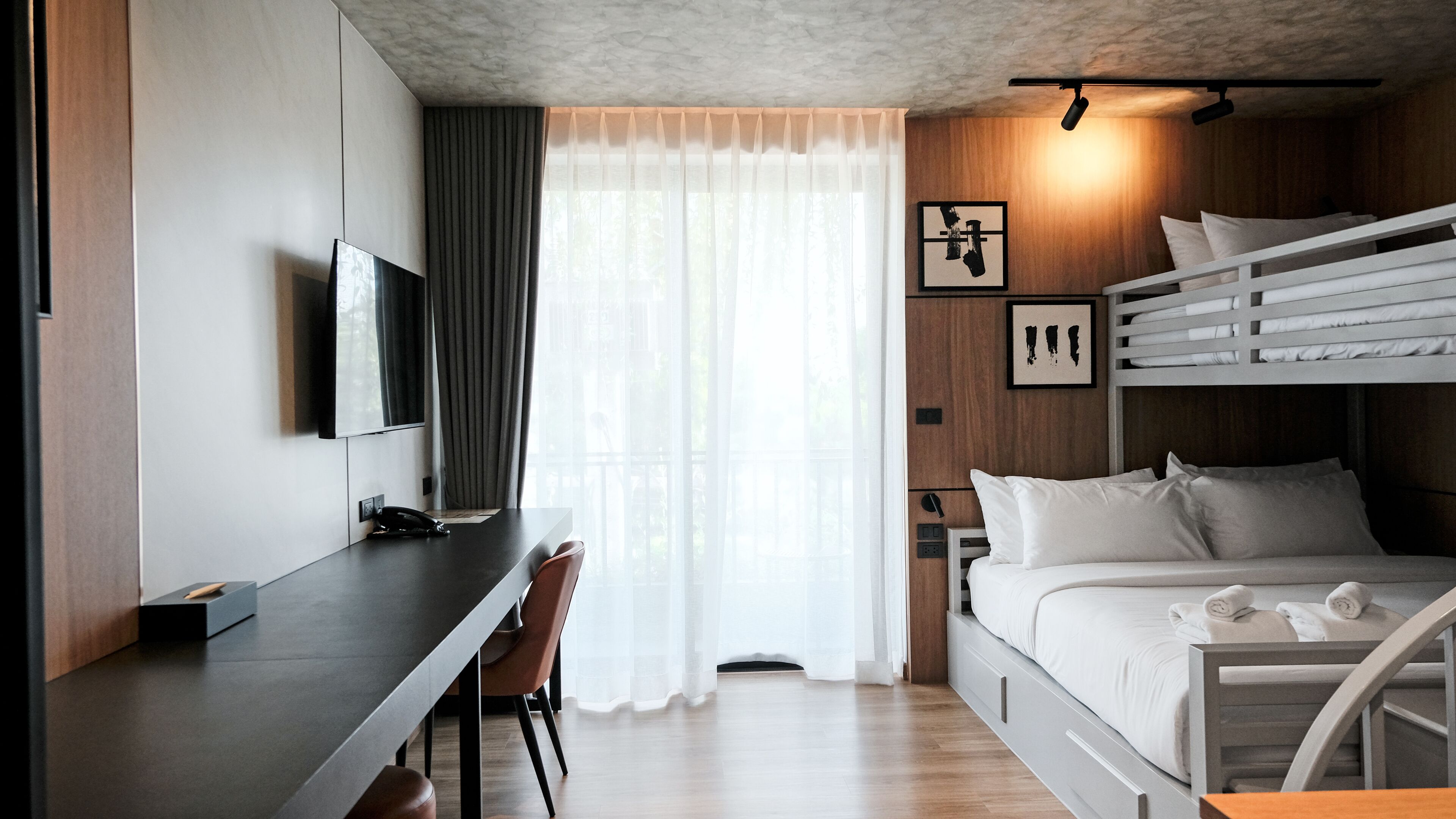 A modern hotel room with a bunk bed, a desk with a chair, a flat-screen TV, and a large window with sheer curtains.
