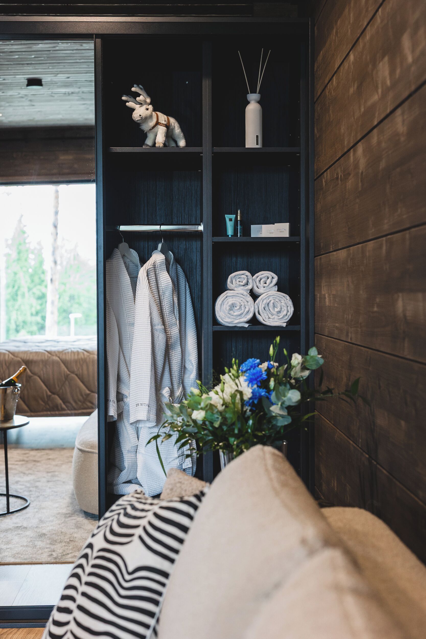 A modern interior with a wooden wall, a black shelving unit, a plush sofa, and a window with a view of trees.