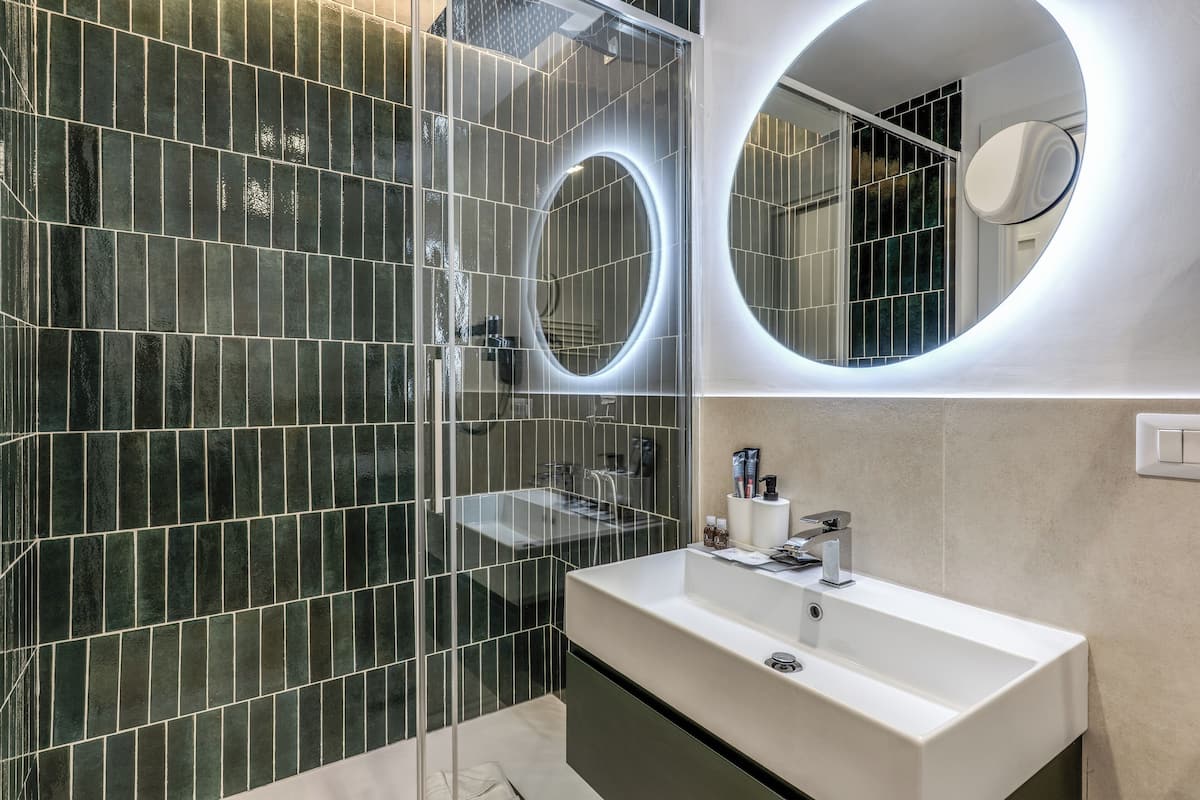 Un baño moderno con ducha de vidrio, lavamanos blanco y espejo redondo con iluminación tipo halo.