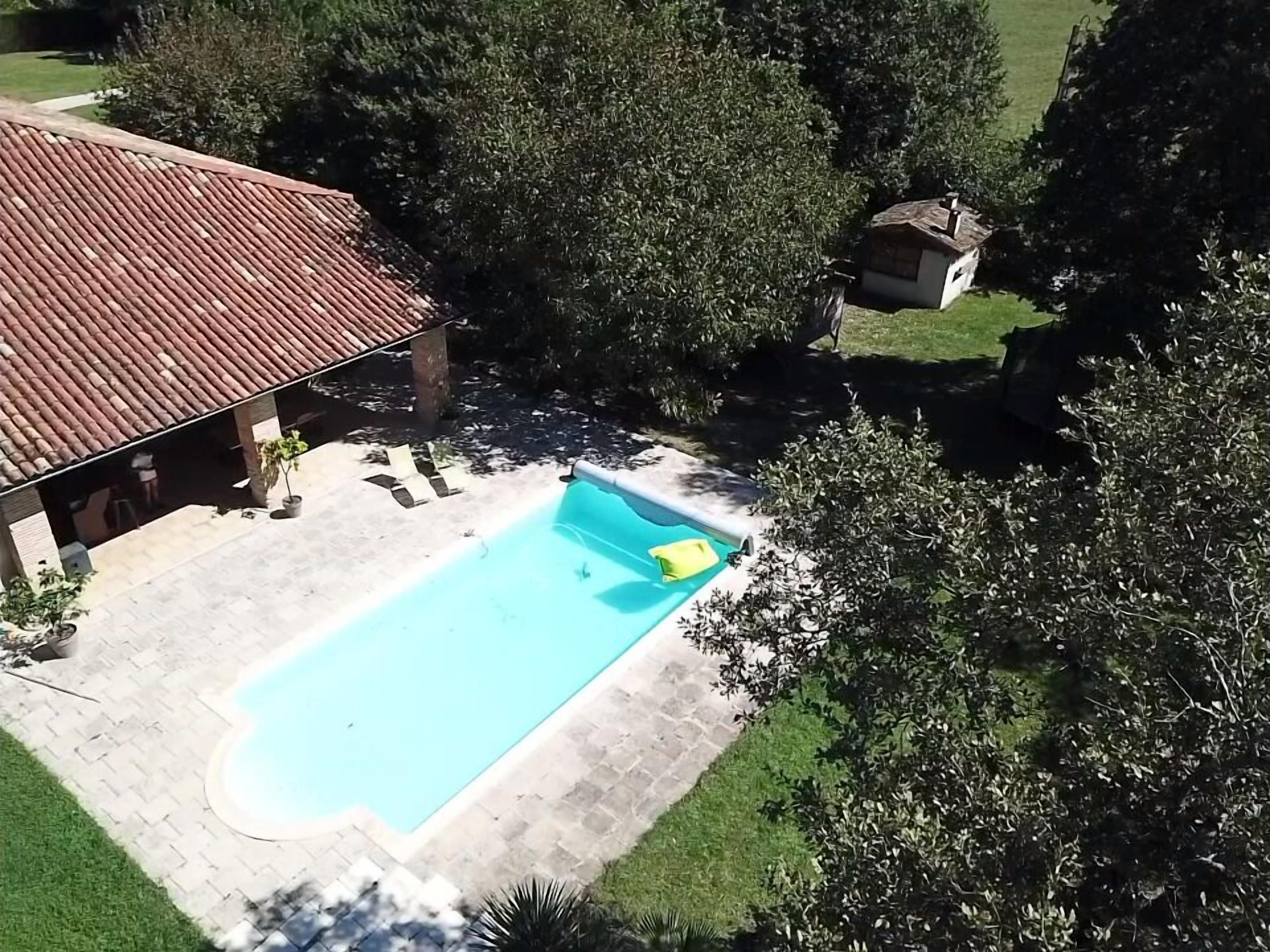 A rectangular swimming pool with a tiled deck, surrounded by trees and a building with a tiled roof.