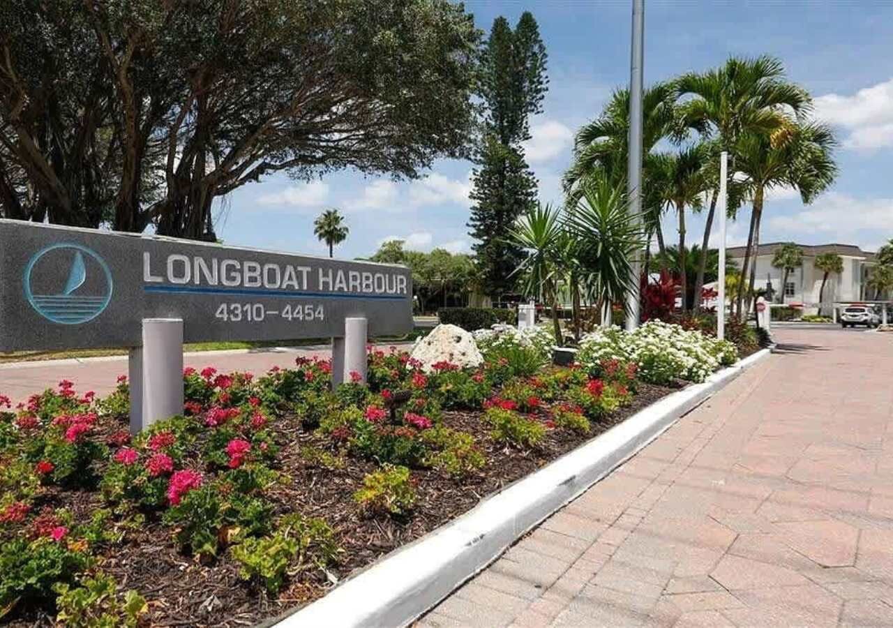 A sign for Longboat Harbour with a contact number, surrounded by a landscaped area with flowers and palm trees.