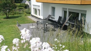 A modern house with a patio area featuring a glass table and chairs, surrounded by a well-maintained lawn and flowering plants.
