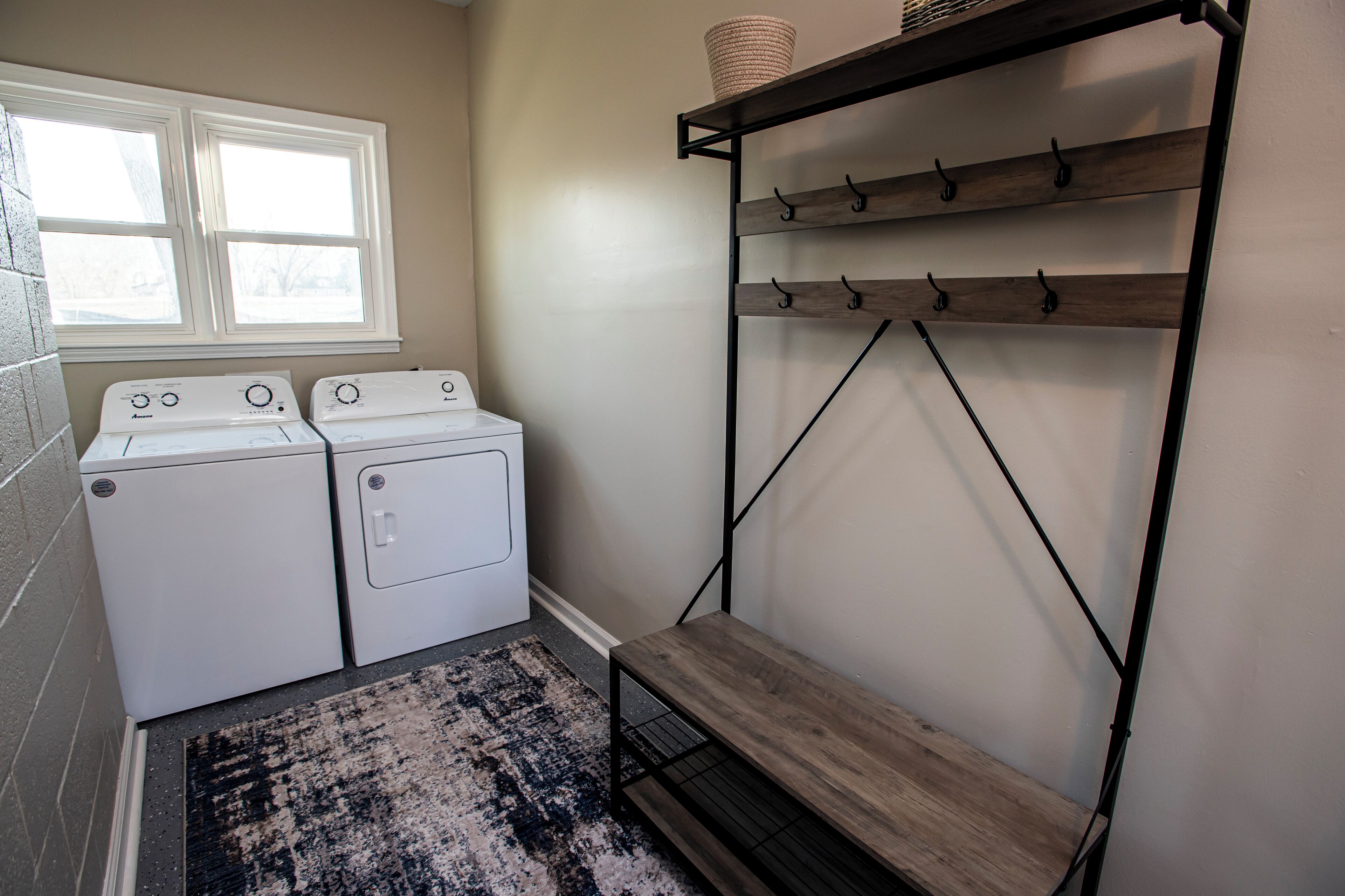 A laundry area with two washing machines, a wooden shelf with hooks, and a rug.