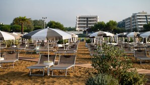 Ein Strand mit Liegestühlen und Sonnenschirmen, feiner Sand und Gebäude im Hintergrund.