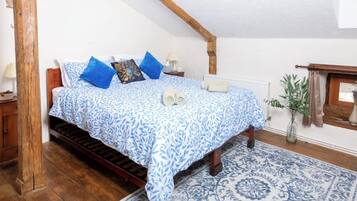Ein Schlafzimmer mit einem hölzernen Bett, einer blauen, gemusterten Tagesdecke, einem blau-weißen Teppich, einem Nachttisch mit Lampe und einer Topfpflanze.